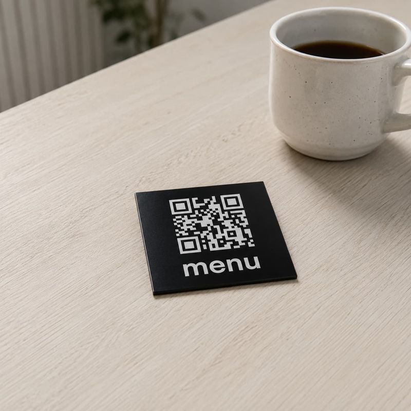 Czarna aluminiowa tabliczka QR z napisem menu na drewnianym stoliku obok filiżanki kawy